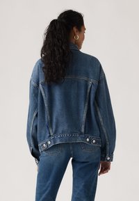 Γυναίκα με μακριά σγουρά σκούρα μαλλιά σε κοτσίδα, που φοράει μπλε τζιν μπουφάν και τζιν παντελόνι, στέκεται γυρισμένη με την πλάτη προς ένα απλό φόντο.