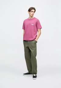 Růžové bavlněné tričko s bílým logem na hrudi, v kombinaci s volnými olivově zelenými kalhotami a černými teniskami. Jednoduchý ležérní design.