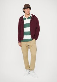 Κεβάλα φούτερ σε μπορντό χρώμα, πολο πράσινο και λευκό ριγέ, μπεζ cargo παντελόνι και λευκά αθλητικά παπούτσια. Καπέλο καφέ με λογότυπο; το μοντέλο στέκεται σε απλό φόντο.