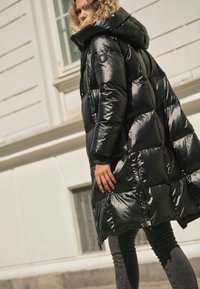 Persoon die een glanzende zwarte donzen jas met capuchon draagt, lopend buiten voor een lichtgekleurd gebouw.