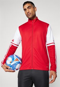 Rode en witte zip-up jack met drie strepen op de mouwen, gecombineerd met een blauw en wit patroon voetbal, gedragen met zwarte broek.