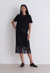 Zwarte outfit met een kort mouwtop met een gestructureerde halslijn en een knie-lange rok met een ingewikkeld patroon, gecombineerd met zwarte, puntige schoenen.