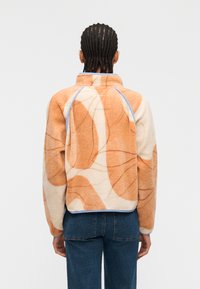 Άτομο με κοτσίδες, φοράει μπεζ και πορτοκαλί φλις μπουφάν με αφηρημένο μοτίβο και μπλε τζιν, στέκεται πλάτη σε λευκό φόντο.