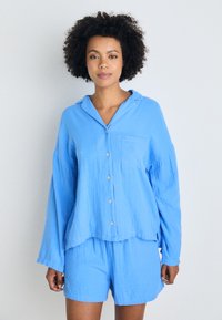 Cămașă bleu deschis, cu nasturi, guler și buzunar pe piept, asociată cu pantaloni scurți asortați. Materialul pare ușor și moale, cu o textură crăpată.