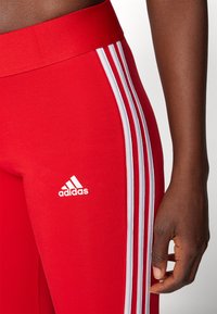 Rdeče športne leggins hlače iz razteznega materiala, ki imajo tri bele črte ob straneh in belo Adidasovo logo na sprednji strani.