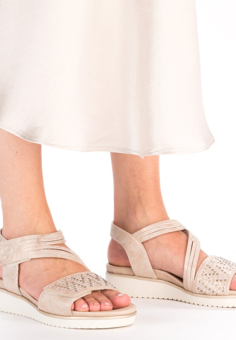 Beige suede sandals feature crisscross straps, embellished upper with studs, and a white platform sole. Soft texture and open design.