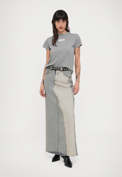 Tetovált nő szürke pólóban, kéttónusú hosszú farmer szoknyában fekete övvel és fekete boka csizmában, sima háttér előtt állva.