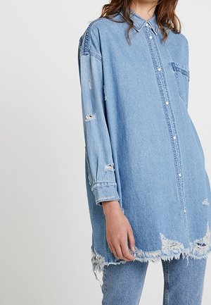 Kobieta ubrana w jasnoniebieską, oversize, przetartą koszulę dżinsową z postrzępionym dołem oraz dopasowane przetarte jeansy, stojąca na jednolitym tle.
