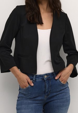 Kvinde iført en sort cropped blazer over en hvid top med blå jeans, hænderne i lommerne, foran en ensfarvet lys baggrund.