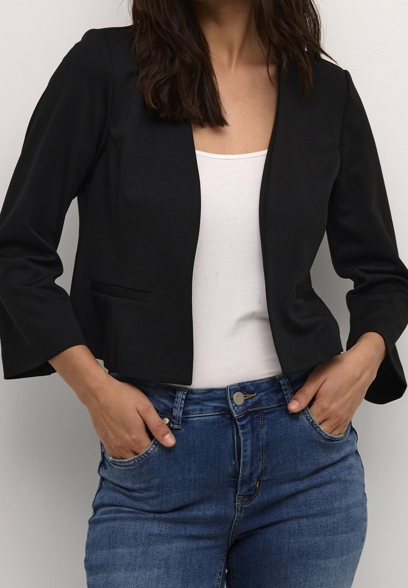 Femme portant un blazer noir court par-dessus un top blanc avec un jean bleu, mains dans les poches, devant un fond clair uni.