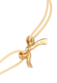 Ciondolo a nastro color oro su una corda beige chiaro. Il ciondolo presenta una finitura liscia con dettagli incisi. La corda è annodata, offrendo possibilità di regolazione.
