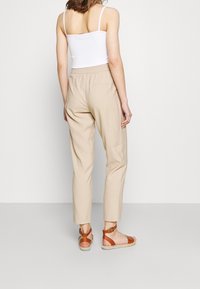 Beige bomullbyxor med elastisk midja, rak skärning och avsmalnande ben. Kombinerade med bruna sandaler med remmar. Modellen vänder sig bort från kameran.