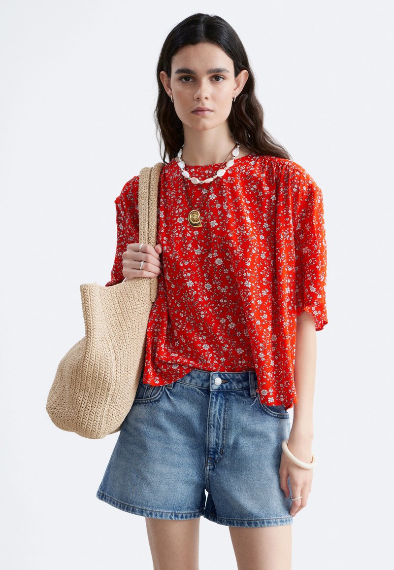 Femme portant un chemisier rouge à manches courtes à motifs floraux et un short en jean, portant un sac beige tissé sur l'épaule.