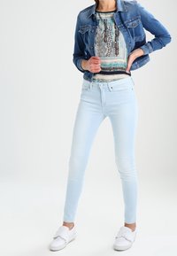 Light blue skinny jeans, a denim jacket, and a patterned top. White slip-on shoes with a flat sole. Smooth texture and casual design.