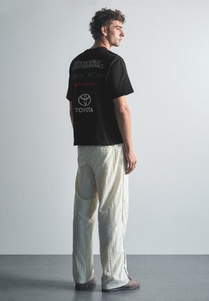 Jeune homme aux cheveux bouclés, debout de profil droit, portant un t-shirt noir Toyota et un pantalon blanc ample sur un fond gris uni.