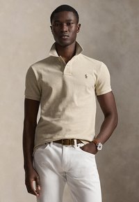 Man draagt een beige poloshirt met opstaande kraag, witte broek, bruine riem en polshorloge, staand tegen een neutrale achtergrond.