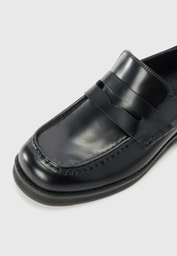 Mocassin en cuir noir à finition lisse, avec un double strap et des coutures renforcées sur les bords. Semelle en caoutchouc plat pour une bonne adhérence.