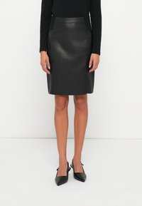 Femme portant une jupe en cuir noire jusqu'au genou, un haut noir à manches longues et des escarpins à talon ouvert noirs à bout pointu, se tenant devant un fond blanc.