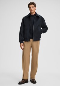 Dark blue, zip-front jacket with a high collar and two front pockets, paired with light brown, wide-leg trousers and dark brown shoes.