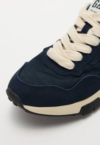 Adidași bleumarin, confecționați din piele întoarsă și materiale textile, cu șireturi crem și o talpă din cauciuc texturat negru. Vârf rotund cu detalii de cusătură.