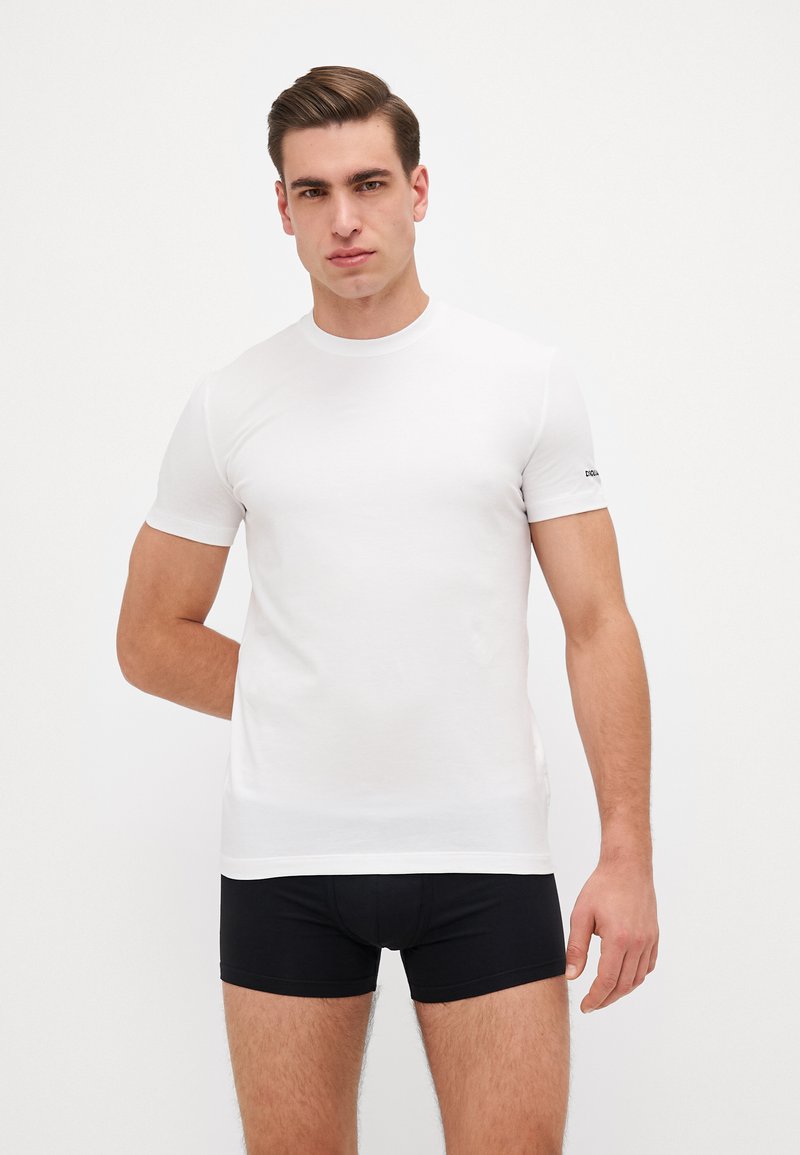 Weißes T-Shirt mit kurzen Ärmeln aus Baumwolle, runder Ausschnitt, mit einem dezenten Markenlogo auf dem linken Ärmel. Kombiniert mit schwarzen, eng anliegenden Shorts.