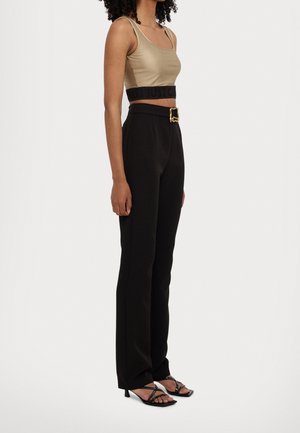 Tan cropped top met zwarte tailleband; zwarte broek met hoge taille en een gouden gesp; zwarte sandalen met bandjes en een vierkante neus.