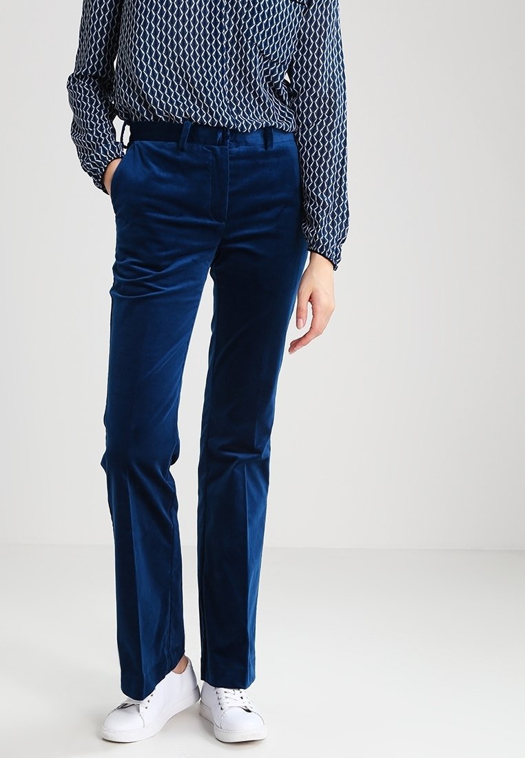 Personne portant un pantalon en velours bleu, des baskets blanches et un chemisier à manches longues à motif, debout avec une main dans la poche.