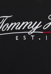 T-shirt nera con il testo ricamato bianco "Tommy" in evidenza, con un accento rosso sotto, su una texture di tessuto liscia.