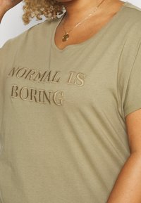 Olivově zelené tričko vyrobené z měkké látky, s vyšívaným textem "NORMAL IS BORING" v kontrastní béžové barvě. Krátké rukávy a kulatý výstřih.