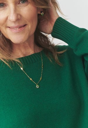 Groene gebreide trui met een geribbelde halslijn, vergezeld van een gouden ketting met een ronde hanger en decoratieve accenten.