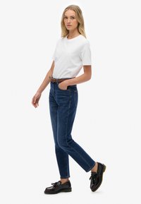 Camiseta blanca de manga corta, vaqueros azules de cintura alta con bolsillos frontales y mocasines negros con borlas. Cinturón trenzado marrón como acento.