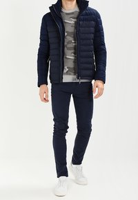 Chaqueta acolchada azul marino con acabado texturizado, suéter gris con camuflaje, pantalones oscuros y zapatillas blancas. Ajuste ceñido y diseño sencillo.