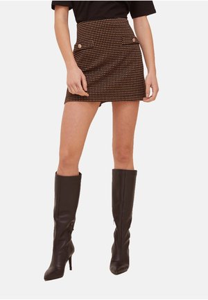 Femme portant une mini-jupe marron à taille haute avec motifs, poches avant, et de grandes bottes en cuir marron foncé à talons.