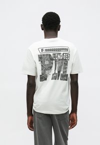 Camiseta branca de algodão com uma estampa gráfica nas costas, apresentando texto e padrões em preto. Corte descontraído, mangas curtas, gola redonda.