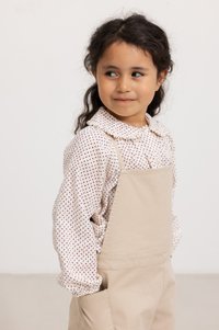 Abito salopette beige con tasche sopra una camicia bianca con motivo che presenta piccoli disegni rossi e blu. Bambino con capelli scuri e ondulati.