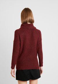 Bordowy sweter dziany z wysokim kołnierzem, prążkowaną fakturą i długimi rękawami, noszony na czarnym ubraniu, prezentujący luźny krój.