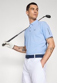 Világoskék poloing fekete CK logóval, fehér nadrággal és tengerészkék övvel. Golfütő a kezében, fehér kesztyűt visel. Bőr látható tetoválásokkal.