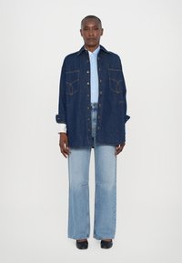 Denim overshirt in dark wash with two front pockets, snap buttons, and contrasting stitching; paired with light blue shirt and flared jeans.