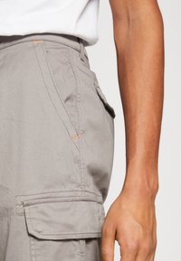 Lichtgrijze cargobroek van zachte stof, met meerdere zakken, contrasterende oranje stikaccenten en een ontspannen pasvorm.