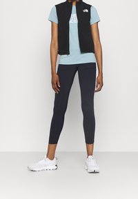 Chaleco negro sin mangas sobre una camiseta azul claro con logo blanco, combinado con leggings oscuros y zapatillas deportivas blancas. Textura de tela suave.