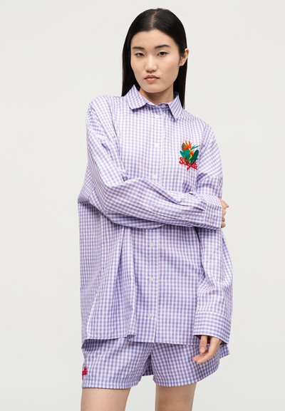 Γυναίκα φοράει υπερμεγέθη πουκάμισο με μοβ καρό κουμπωτό και ταιριαστό σορτς με κεντήματα λουλουδιών στο στήθος και στα σορτς, στέκεται μπροστά σε λευκό φόντο.