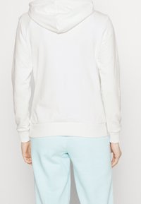 Sudadera blanca con capucha, hecha de una tela suave y texturizada. Presenta un dobladillo y puños de canalé, combinada con pantalones azul claros para un look casual.