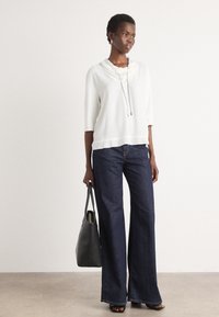 Witte top met kraag en korte mouwen, gecombineerd met donkerblauwe wijde jeans. Model houdt een zwarte tote bag vast. Soepele texturen, minimalistisch ontwerp.