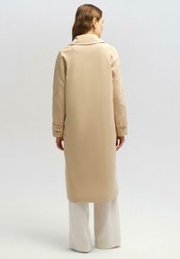 Palton lung bej cu guler, având accente decorative pe mâneci și manșete cu nasturi. Purtat peste pantaloni albi cu croi larg.