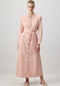 Dlouhé růžové šaty z lehké tkaniny, s knoflíkovou přední částí se modrými knoflíky, dlouhými rukávy a páskem v pase, který se zavazuje.
