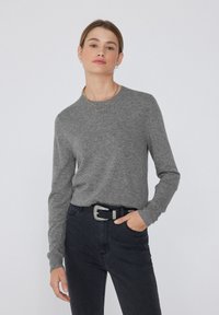 Frau mit hellbraunen Haaren, die zu einem Pferdeschwanz gebunden sind, trägt einen grauen langärmligen Pullover, schwarze Jeans mit einem Gürtel mit silberner Schnalle und posiert mit einer Hand in der Tasche.