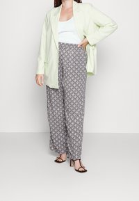 Vrouw in een lichtgroene blazer, witte top, geprinte broek met wijde pijpen en zwarte hakken met bandjes, staand tegen een effen lichte achtergrond.