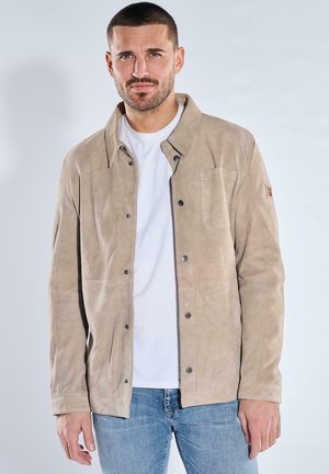 Mann trägt beige Wildlederjacke mit Knöpfen über weißem Hemd und blauer Jeans, steht vor einem einfarbigen hellen Hintergrund.