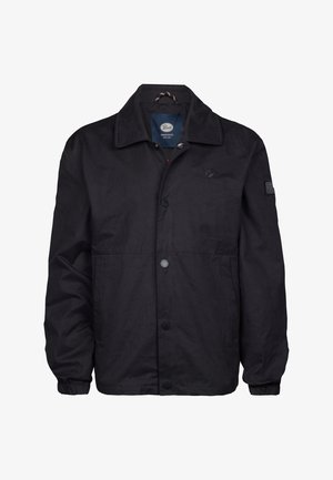 Czarna, lekka kurtka z zatrzaskami, beżowa koszula, oliwkowe spodnie i białe trampki; chłopiec stoi uśmiechnięty na tle jednolitym.