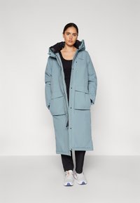 Cappotto lungo e leggero di colore blu chiaro imbottito con cappuccio, presenta fodera nera, bottoni a pressione e tasche frontali. Indossato sopra una maglietta nera e pantaloni neri.